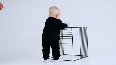 lovely baby boy in black suit stands at the metal grate cube. man comes up to child trying to turn him to camera. white backdrop.