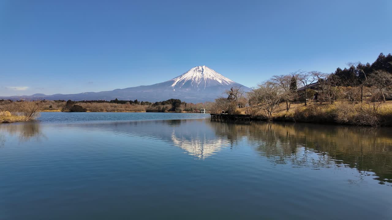 Un'ampia inquadratura serena del Monte Fuji innevato, riflesso nelle acque calme del Lago Tanuki sotto un cielo azzurro e limpido. Perfetto per contenuti di viaggio e natura.