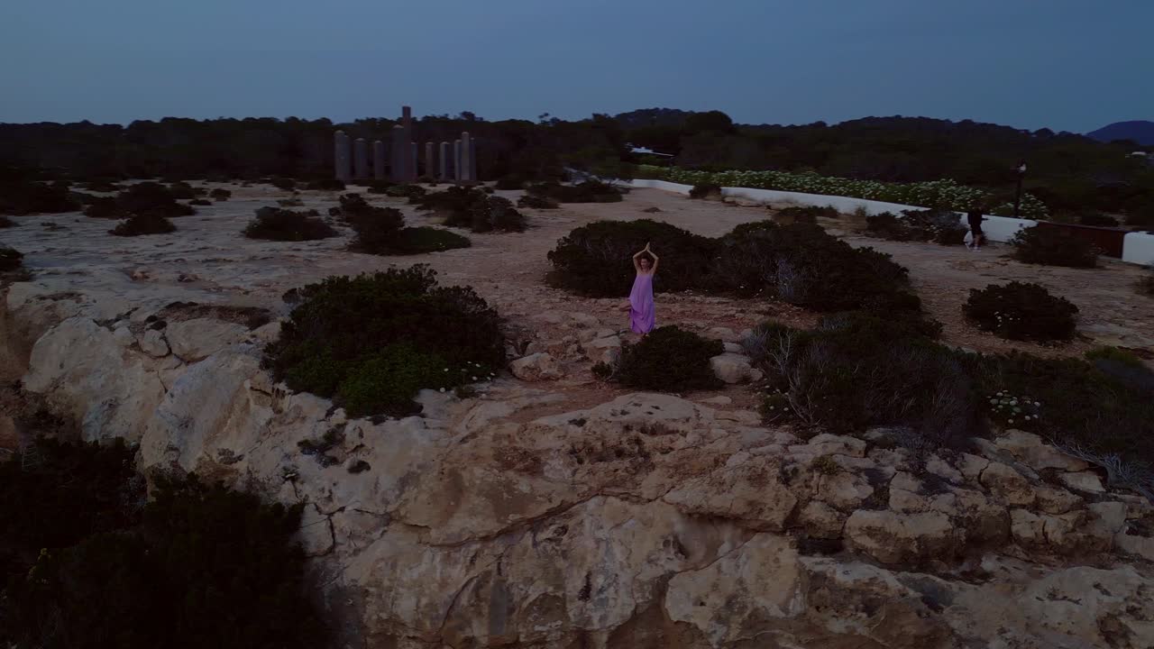 Woman meditating in yoga position on a cliff overlooking the sea at sunset in Ibiza. Perfect aerial view flight wide orbit overview drone