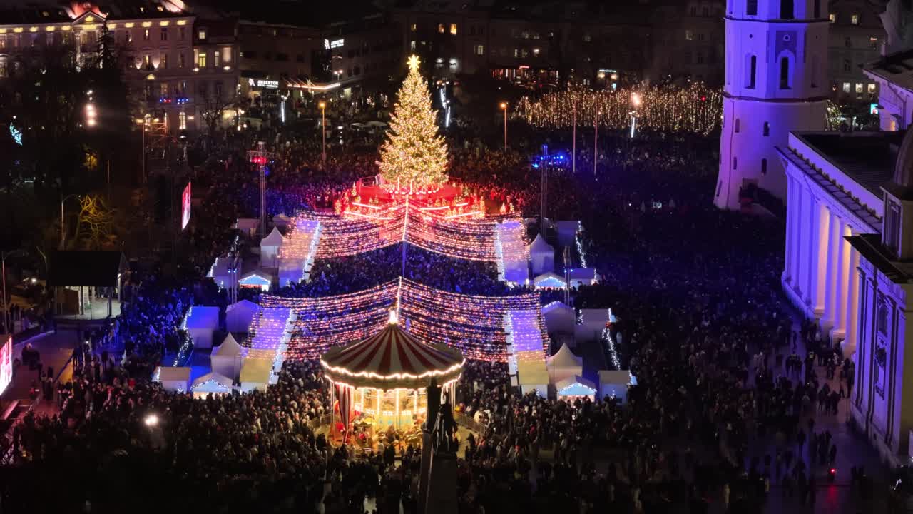 Crowds gather around a towering Christmas tree and carousel under bright holiday lights, creating a lively festive atmosphere filled with colour, movement, and winter celebration joy
