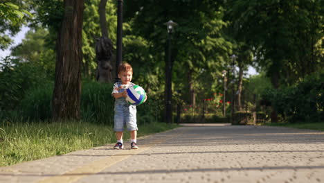 active caucasian baby boy playing with a ball in the park. kid throws the toy and runs happily by the road.