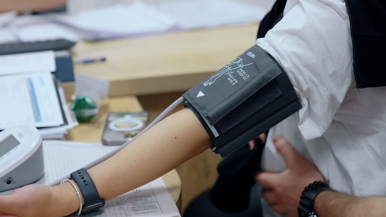 Measuring blood pressure to a pregnant woman. Unrecognized expecting lady sits with a device belt on her arm and her husband's hand strokes her belly.