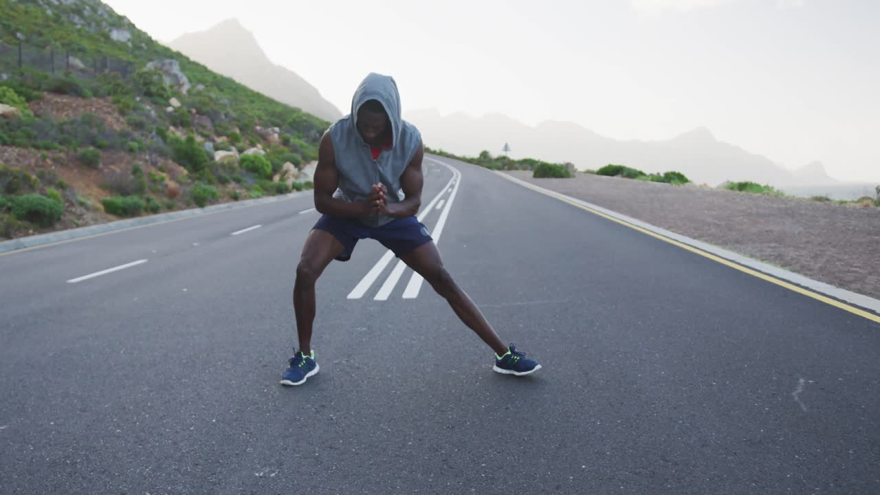 African american man wearing a hoodie performing stretching exercise on the road