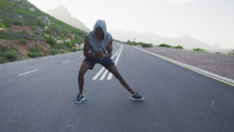 hombre afroamericano con capucha realizando ejercicios de estiramiento en la carretera
