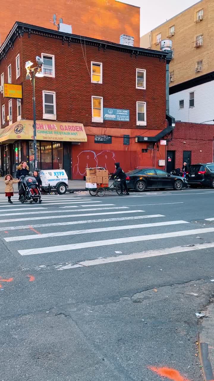New York, USA, 11 May 2025: People in traditional attire crossing urban street near brick buildings. Man in black hat and woman in puffer coat walking on asphalt road near white truck