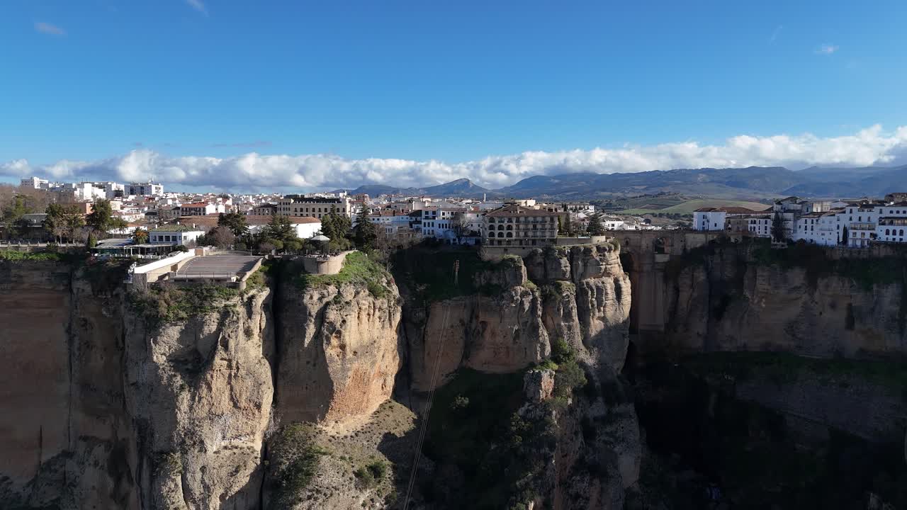 Aerial view of Ronda, Spain, highlighting the stunning Puente Nuevo bridge on dramatic cliffs, with a picturesque cityscape under a bright blue sky and fluffy clouds