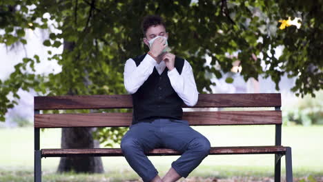 an elegant young man in a park wearing a black waistcoat and white shirt putting on a green protective covid-19 mask, sitting on a park bench and casually relaxing before standing up, static 4k shot