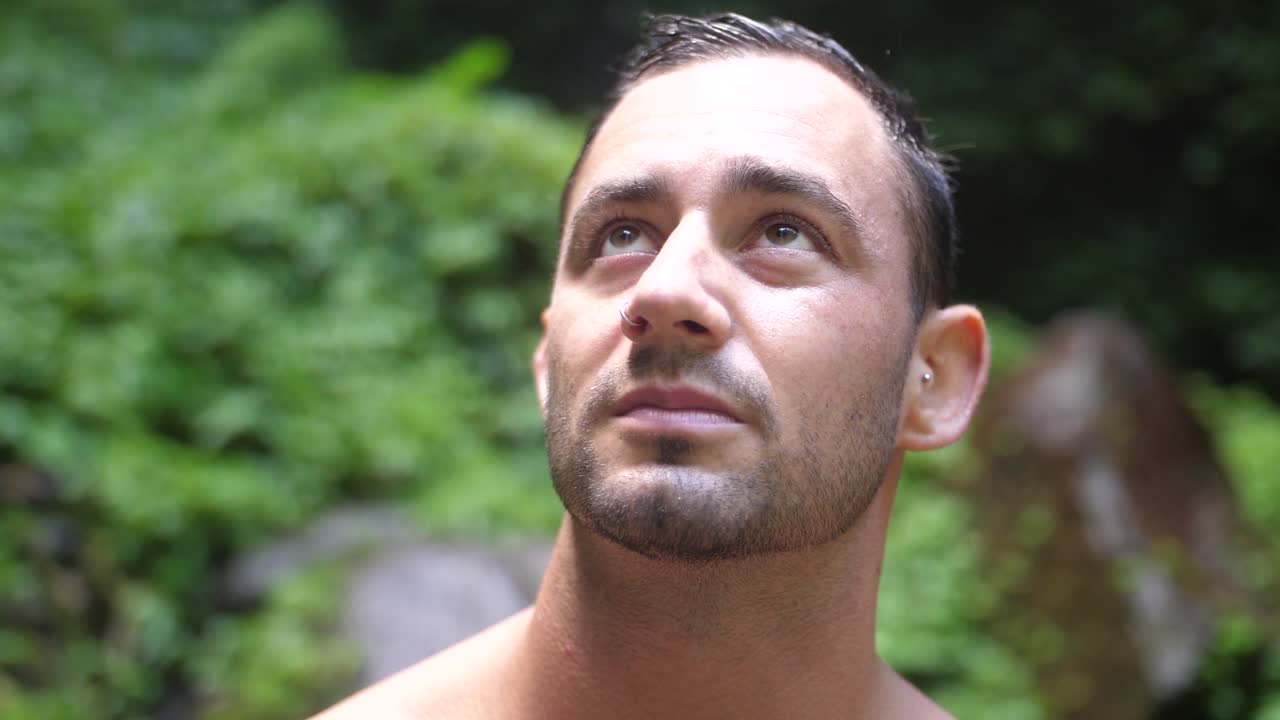 진지한 얼굴 표정을 가진 젊고 매력적인 남자가 카메라를 바라보고 하늘을 향하고 발리의 무성한 녹색 정글에서 주변 환경을 바라보고 있습니다.