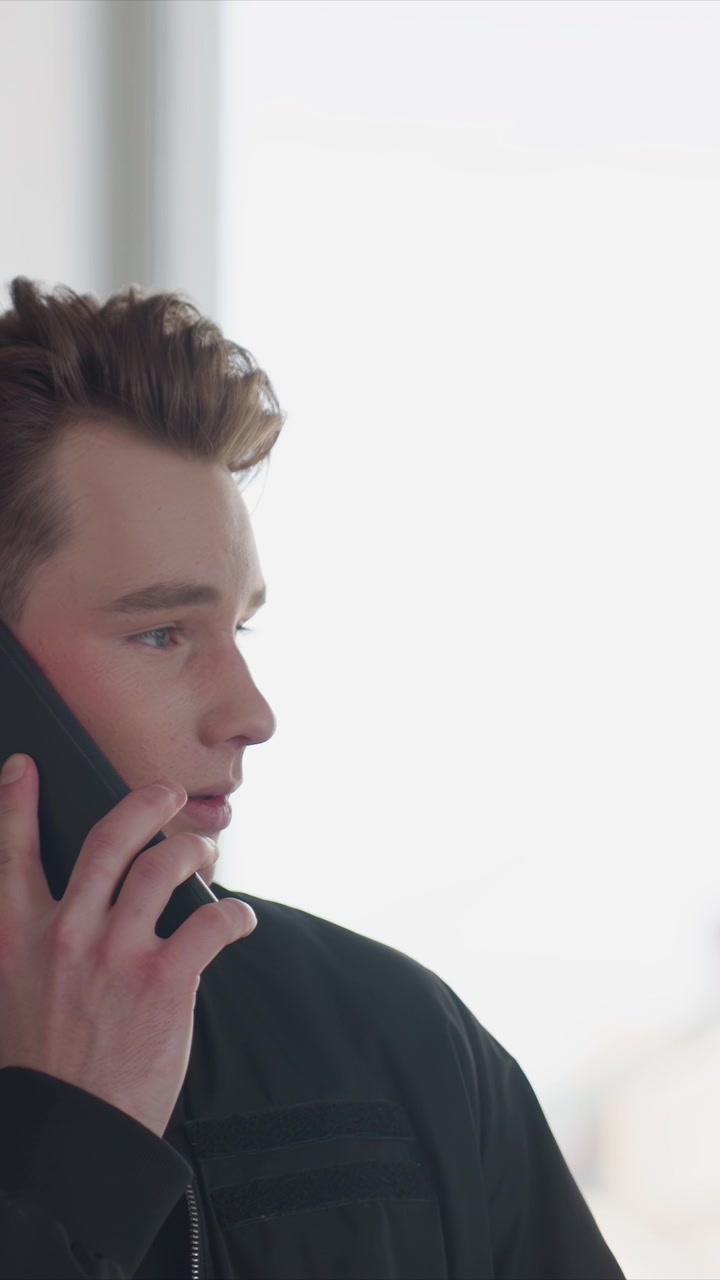 Close up of young man talking on his smartphone indoors. Man holds device to his ear while wearing black jacket. Scene remains consistent throughout focusing on head and upper body