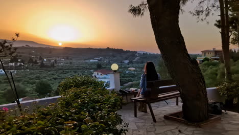 young woman enjoys mountain sunset while sitting on bench, back view