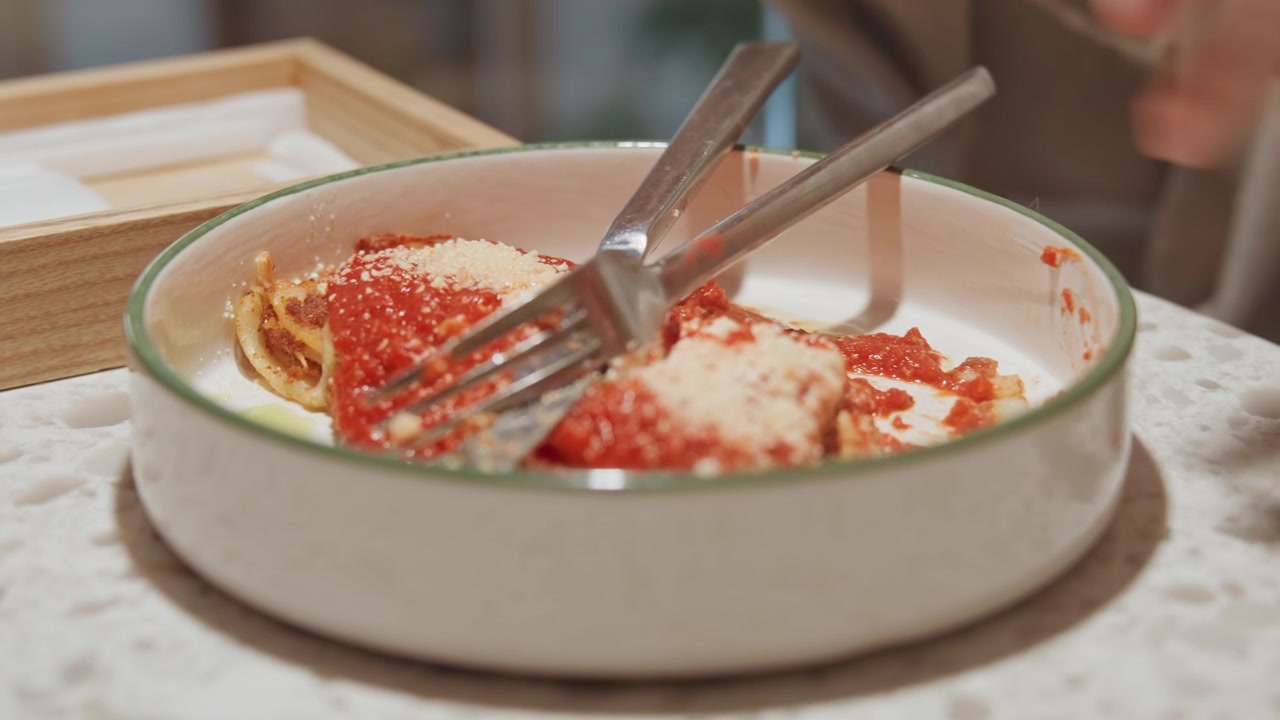 Close up of person using fork and knife to cut into red sauce covered food in white dish. Fork knife utensils interact with meal on speckled countertop near wooden tray. Soft lighting
