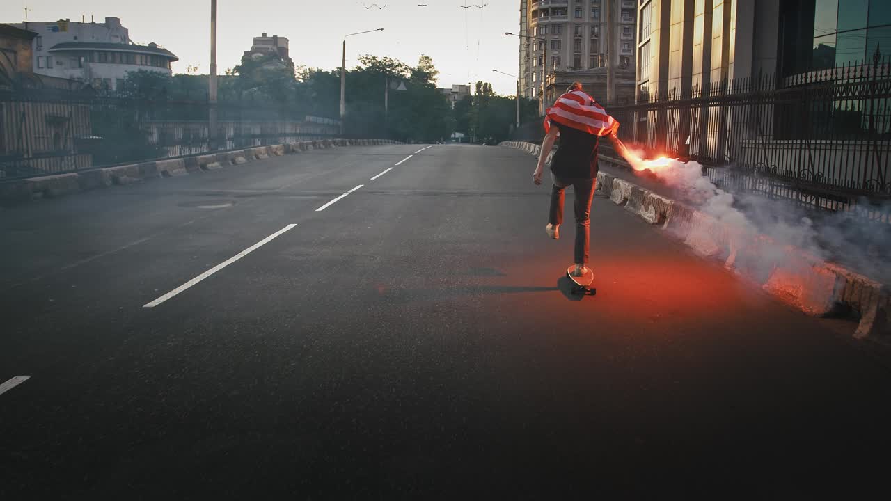 un joven con una bandera de los estados unidos atada en el cuello está montando en patineta a lo largo de una calle desierta con una llamarada de señal roja en la mano.