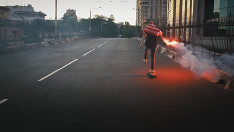 young man in flag of usa tied on his neck is riding skateboard along deserted street with burning red signal flare in hand. slow motion, back view