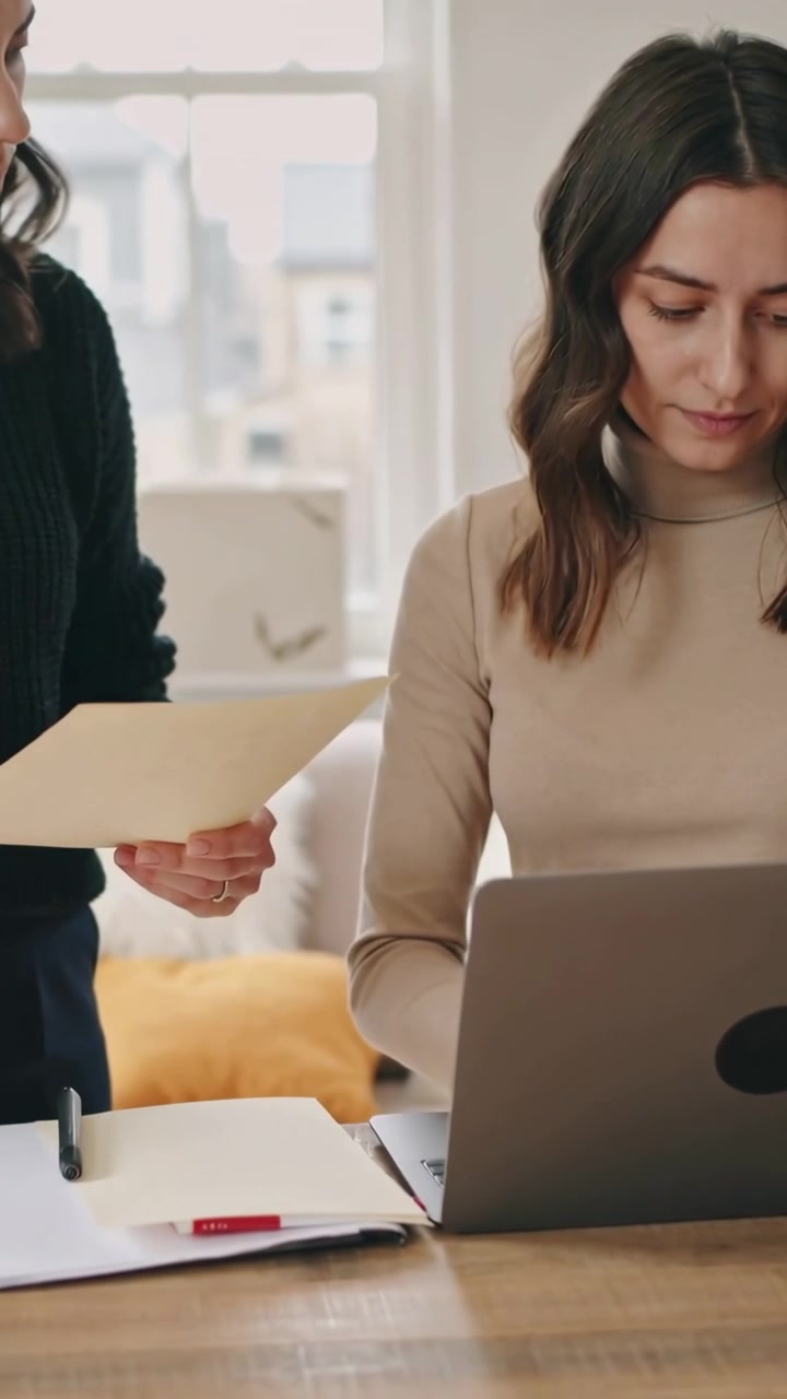 Video verticale: Donna che digita al laptop al tavolo vicino alla finestra, collega che mostra un documento per la revisione