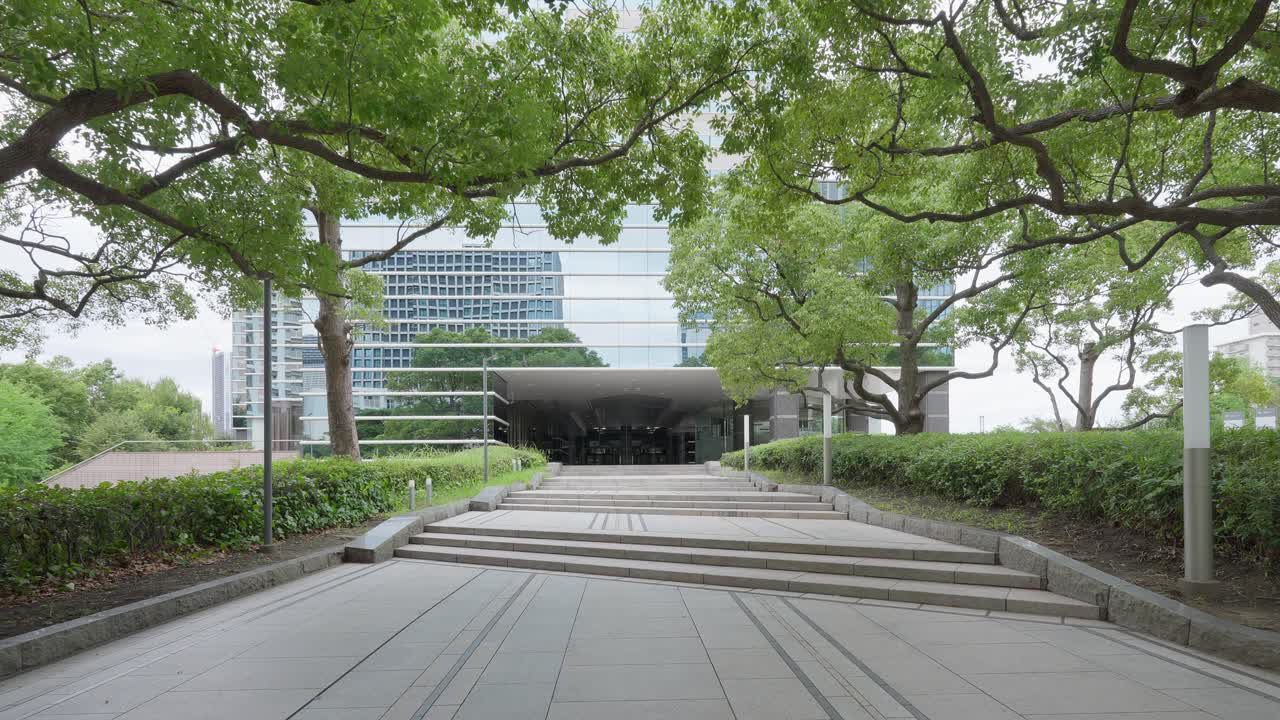 A peaceful shot of a paved walkway and stairs leading toward a modern building, surrounded by green trees and foliage
