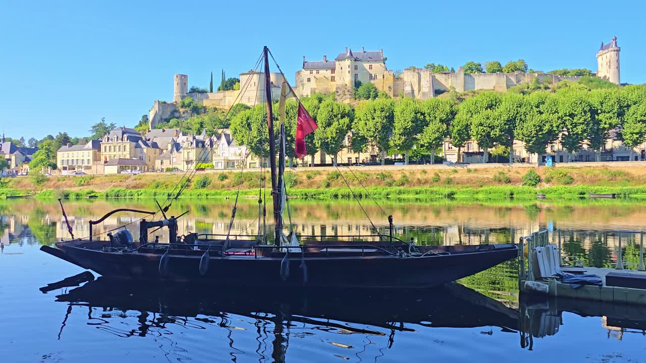 A static shot shows a boat in shadow moored on the river, with the medieval fortress of Chinon and the town visible in the background under a clear blue sky