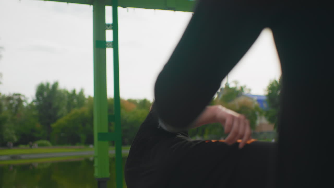 Young man blocking high kick near lakeside pavilion, foreground leg creates dramatic silhouette across frame, calm expression after successful block, sunlit green beams and water reflection add