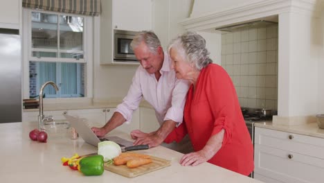 Feliz-Pareja-De-Ancianos-Caucásicos-En-La-Cocina-Usando-Una-Computadora-Portátil-Para-Preparar-La-Receta-Antes-De-Preparar-La-Comida