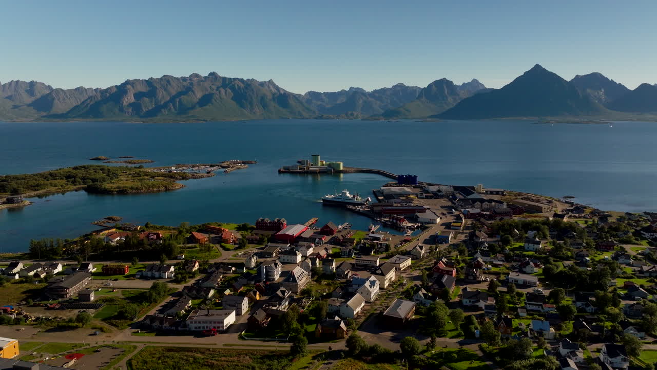 Aerial drone view of Melbu town in Hadsel, Norway. Features the harbor with a ferry, colorful houses, and majestic Lofoten mountains under a blue sky