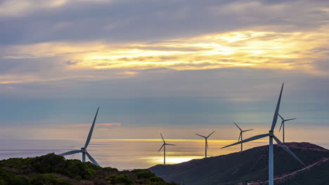 turbinas eólicas girando en el viento generando electricidad con una puesta de sol retroiluminada de cielos naranjas y nubes en movimiento sobre el paisaje y el océano