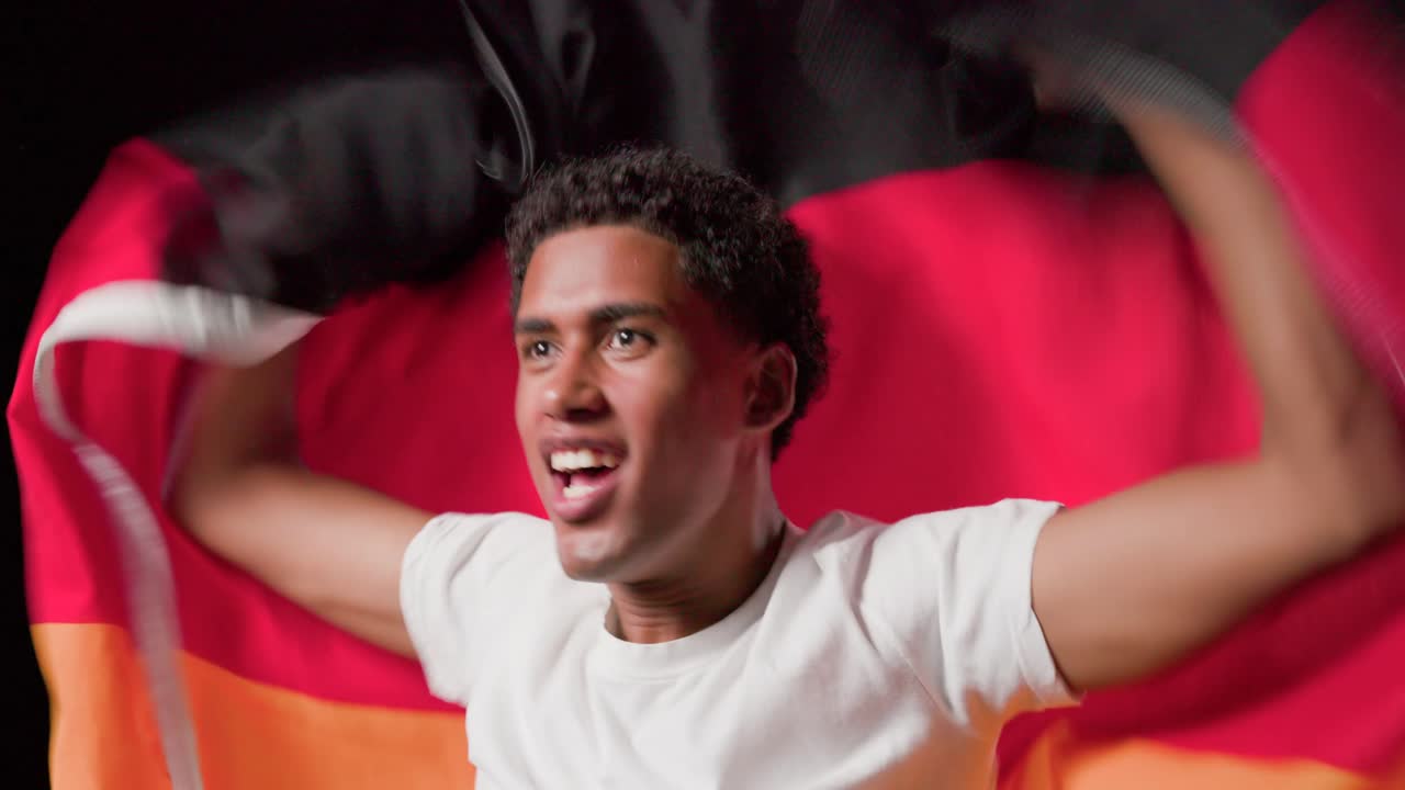 Sonriendo y hablando, un hombre se abre la chaqueta para revelar una bandera roja y naranja mientras celebra en un estudio.