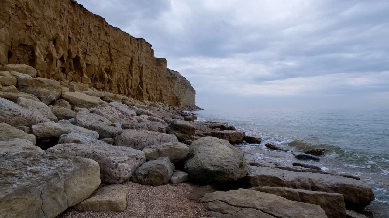 Slow motion of a Jurassic Coast cliff in Dorset from top on the beach with gentle water running up against large rocks and boulders. Sheer face golden cliffs captured on a cloudy evening.