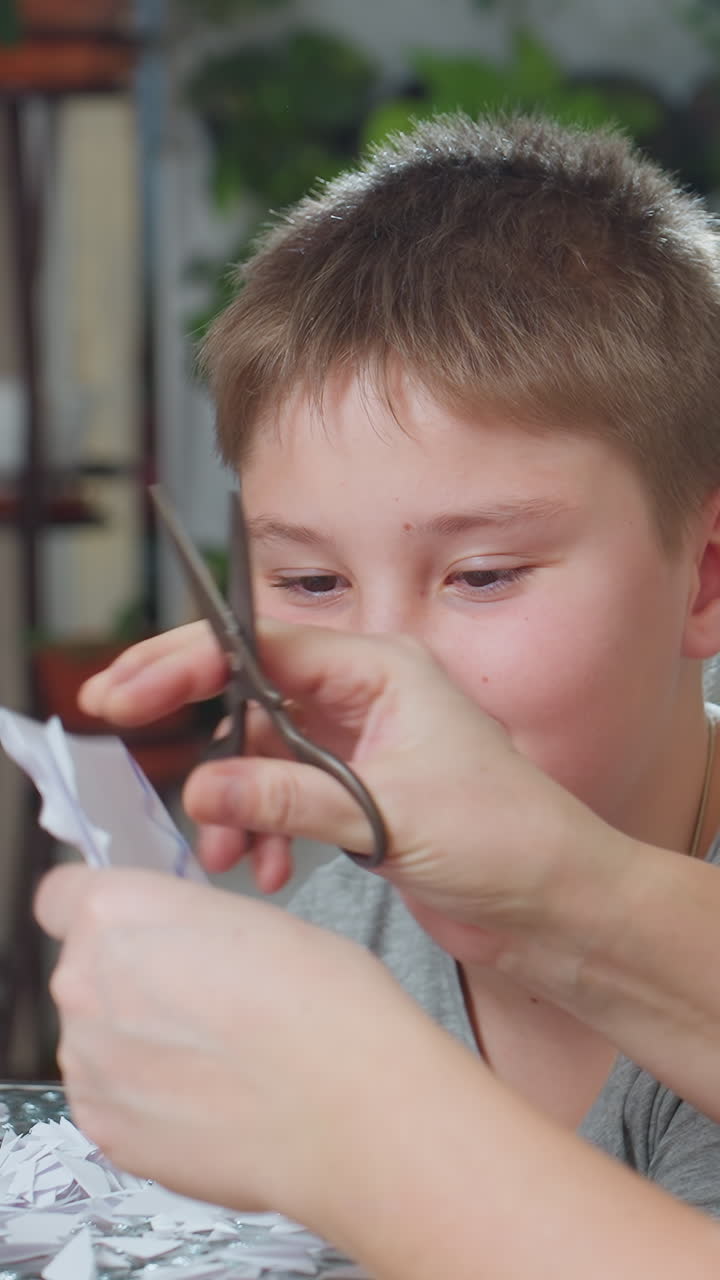 Niño caucásico sonriente observando cómo unas tijeras cortan papel, adulto recortando un patrón de copo de nieve, acogedora escena de manualidades en interior, expresión de entusiasmo, planta decorativa de fondo, actividad familiar práctica