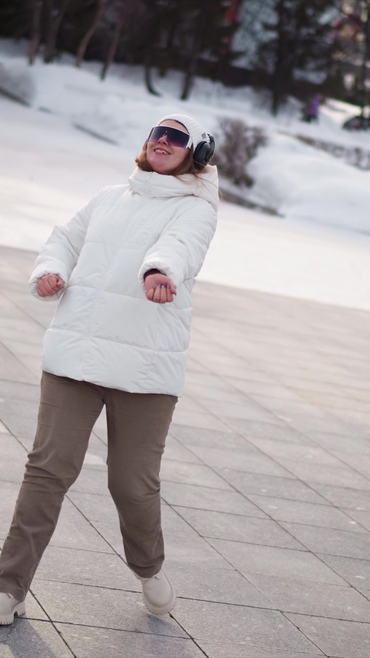 Woman dances energetically on paved area with snow covered ground in background. Woman wears white puffer jacket beige pants and headphones moving her arms and legs in various dance poses