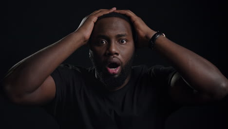 stressed afro guy posing on black background. guy holding hands on head indoors