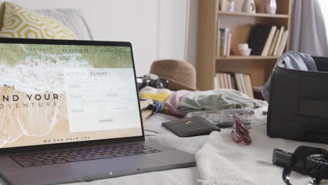 open suitcase on bed at home being packed for summer holiday with laptop showing booking details of flights and hotel