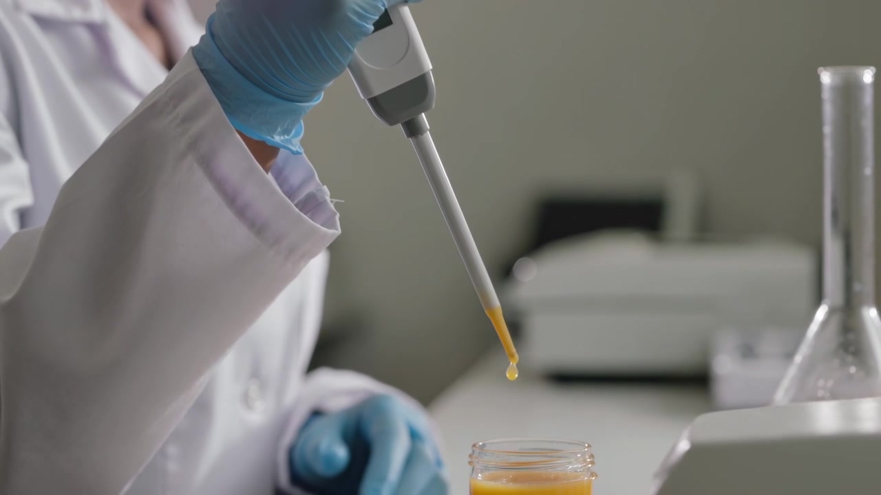 Pressing pipette, lab tech in white coat blue-gloved dispensing orange droplet into jar for assay