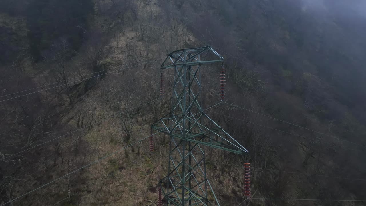 High voltage pylon and power lines above a forest. Drone descends while tilting up, revealing a misty mountain and cloudy sky in rainy weather. Western Norway