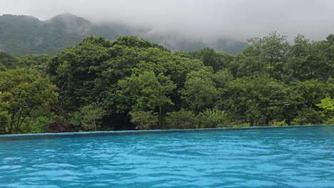 Edge-of-empty-infinity-pool-overlooking-gorgeous-lush-green-trees-mountain-slopes-on-a-rainy-day