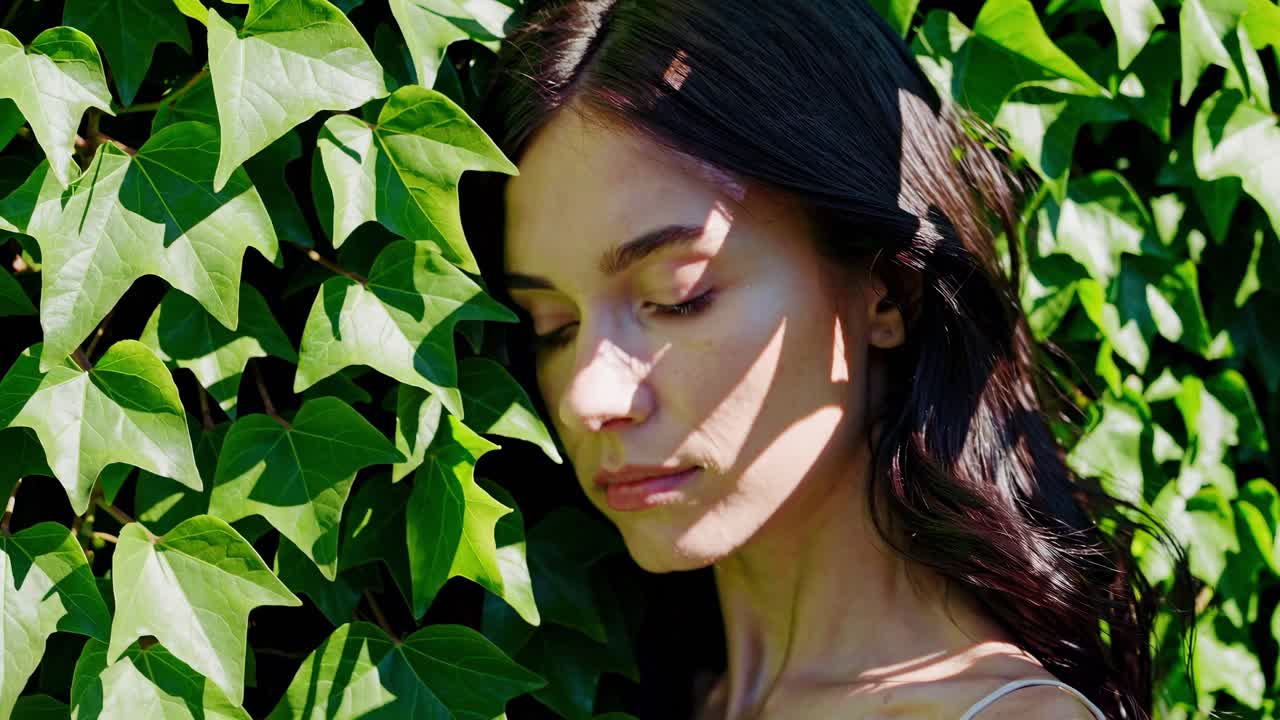 Pensive young woman with long dark hair posing near an ivy hedge in a sunny day, she has leaf shadows projected on her face and neck and is wearing a light colored top