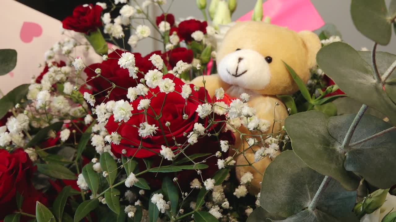 a soft teddy bear nestled among vibrant red roses, white baby's breath, and green foliage. Romantic floral arrangement with a warm, affectionate mood