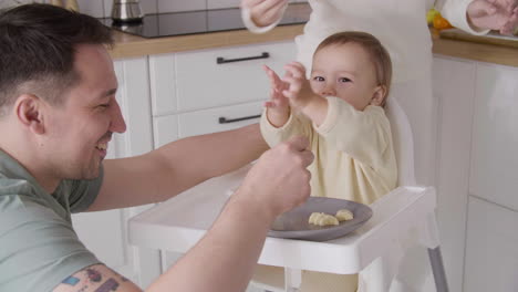 Vater-Füttert-Sein-Glückliches-Kleines-Mädchen,-Das-In-Ihrem-Hochstuhl-In-Der-Küche-Sitzt,-Während-Mutter-Avocadoscheiben-Auf-Den-Teller-Legt