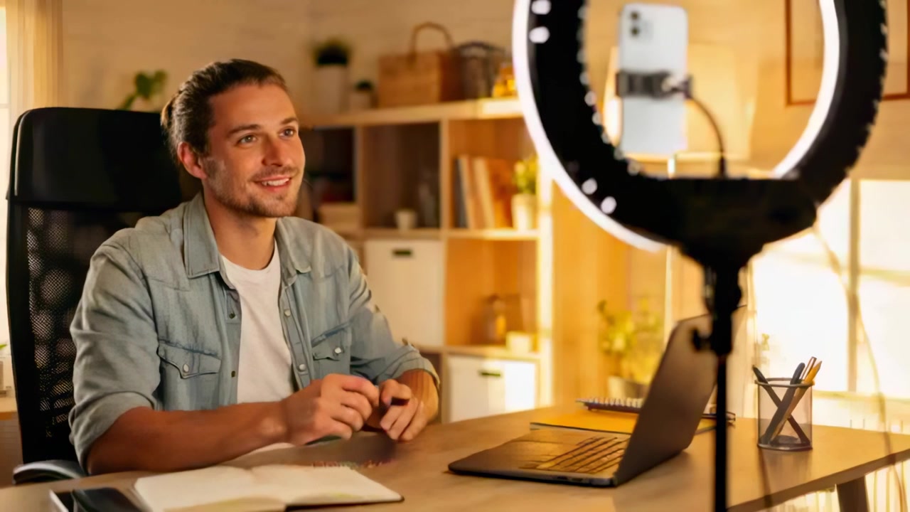 Hombre sonriendo frente al teléfono dentro de un aro de luz, con camisa vaquera saludando, transmitiendo desde la oficina en casa.