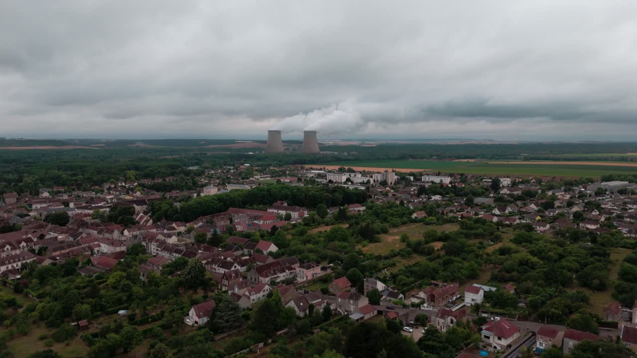 Static high-angle aerial drone shot of a French town with a nuclear power plant on the horizon. Steam rises from the cooling towers against a cloudy, overcast sky