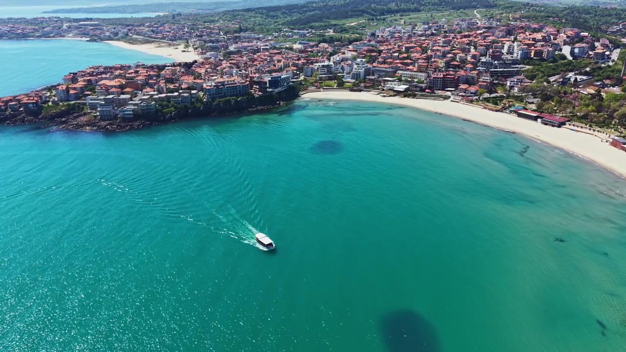 Vista costiera di una piccola barca che naviga in acque limpide vicino a una spiaggia e una città con edifici sullo sfondo a mezzogiorno.