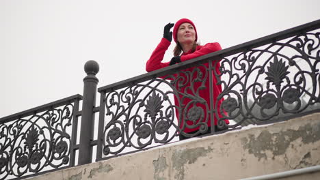 mujer con gorra roja y capucha descansa las manos en una intrincada barandilla de hierro bajo un cielo de invierno brillante, sonriendo con expresión serena y colocando la mano derecha en la cabeza mientras mira hacia la distancia