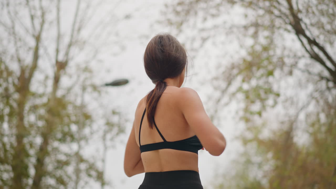 Close-up view of lady swinging her body under bright sunlight, outdoors in park with dry trees in the background, showing fitness exercise, stretching, and physical activity