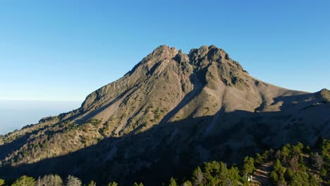 drone ascends above pine forest, framing the steep rocky summit of nevado de colima under a clear blue sky