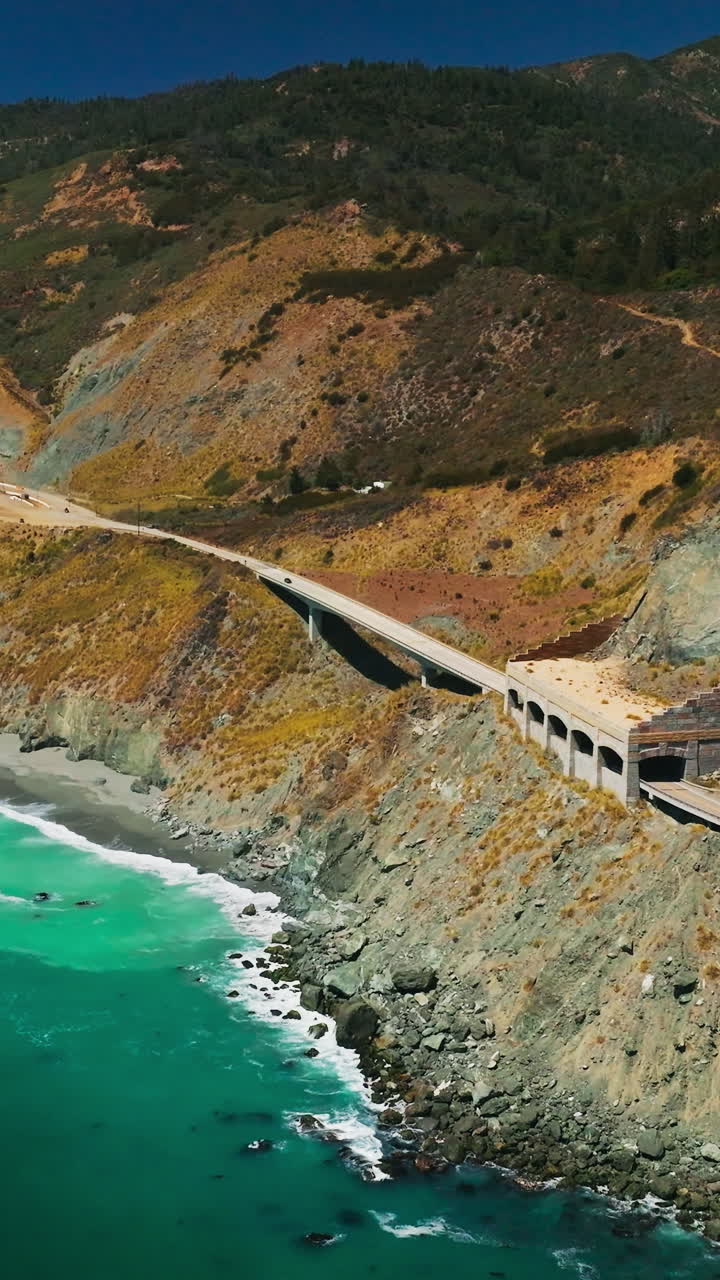 Idyllic scenery of turquoise ocean and crushing waves at the rocks. Highway passing through the mountains. Sunny day backdrop. Vertical video