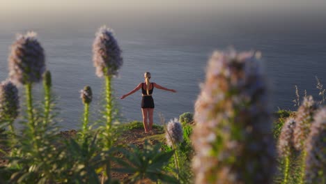 woman raising arms meditating on cliff overlooking sea, pride of madeira flower bokeh