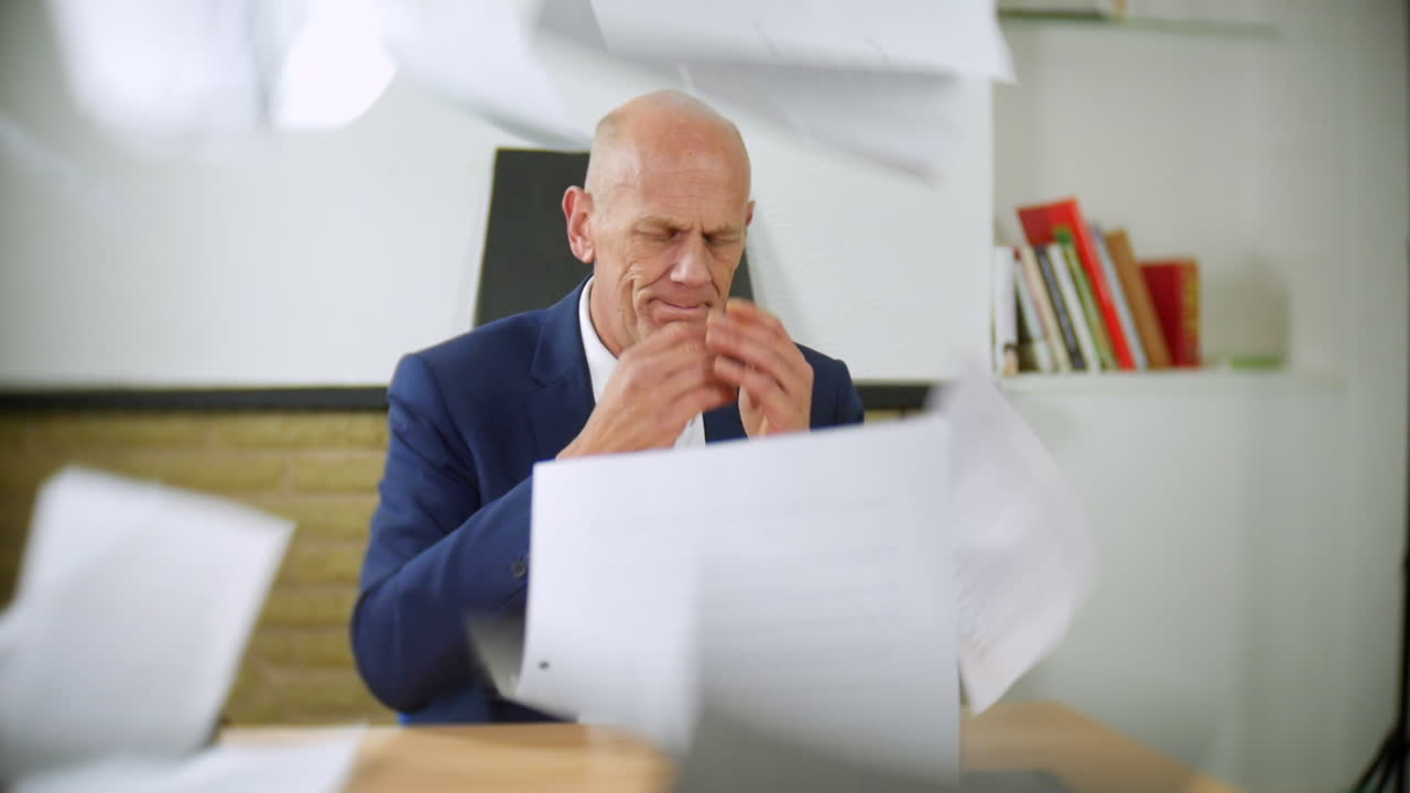 An angry businessman sitting at a desk throws papers in the air