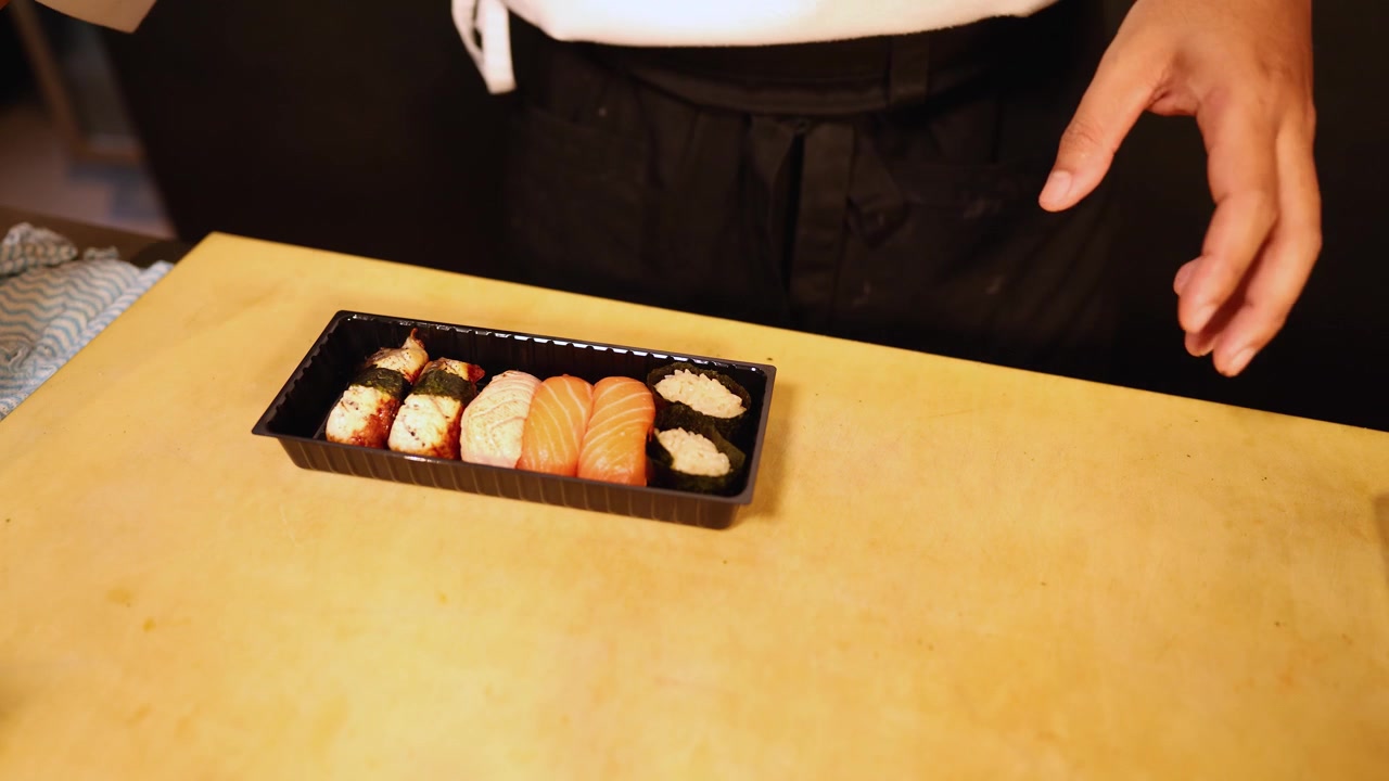 シェフが木製カウンターの上で新鮮なサーモンとウナギの握りをプラスチック製デリバリー容器に入れる