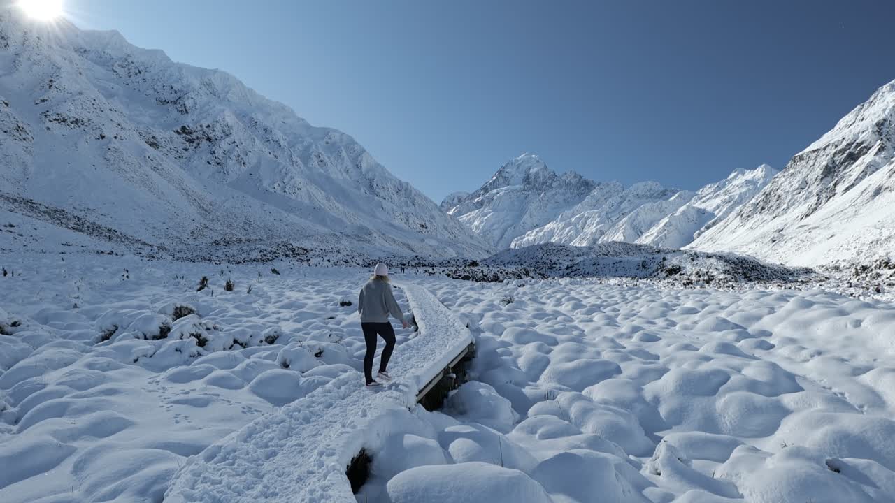 A young woman walks alone along the wooden footpath of the snow-blanketed Hooker Valley Track, surrounded by alpine peaks under clear blue skies