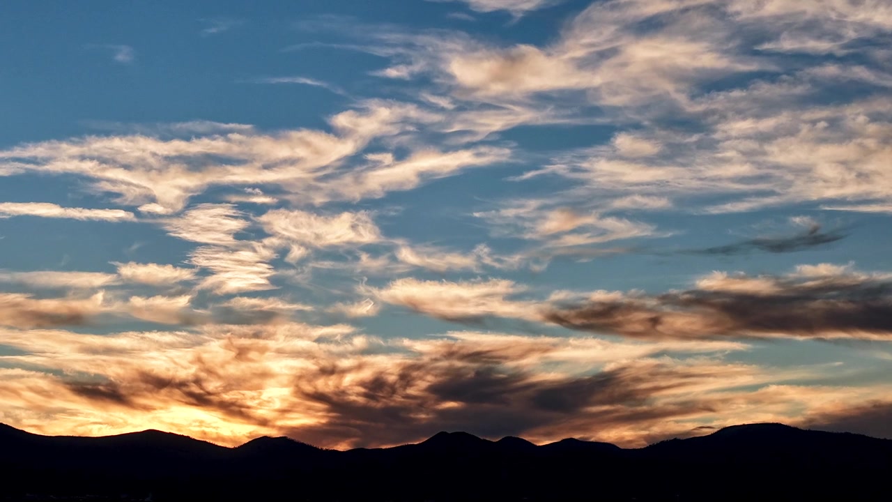 4K UHD timelapse capturing the luminous afterglow of a Southwestern twilight. Wispy, highly textured clouds glow with warm orange hues fading into blue above a stark black mountain silhouette.