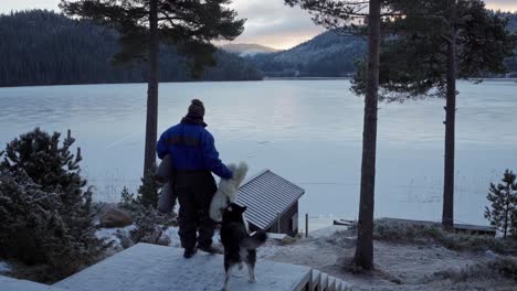 rear view of man with dog walking down the stairs carrying mat and fur rug overlooking frozen lake in forest hills near trondheim, norway