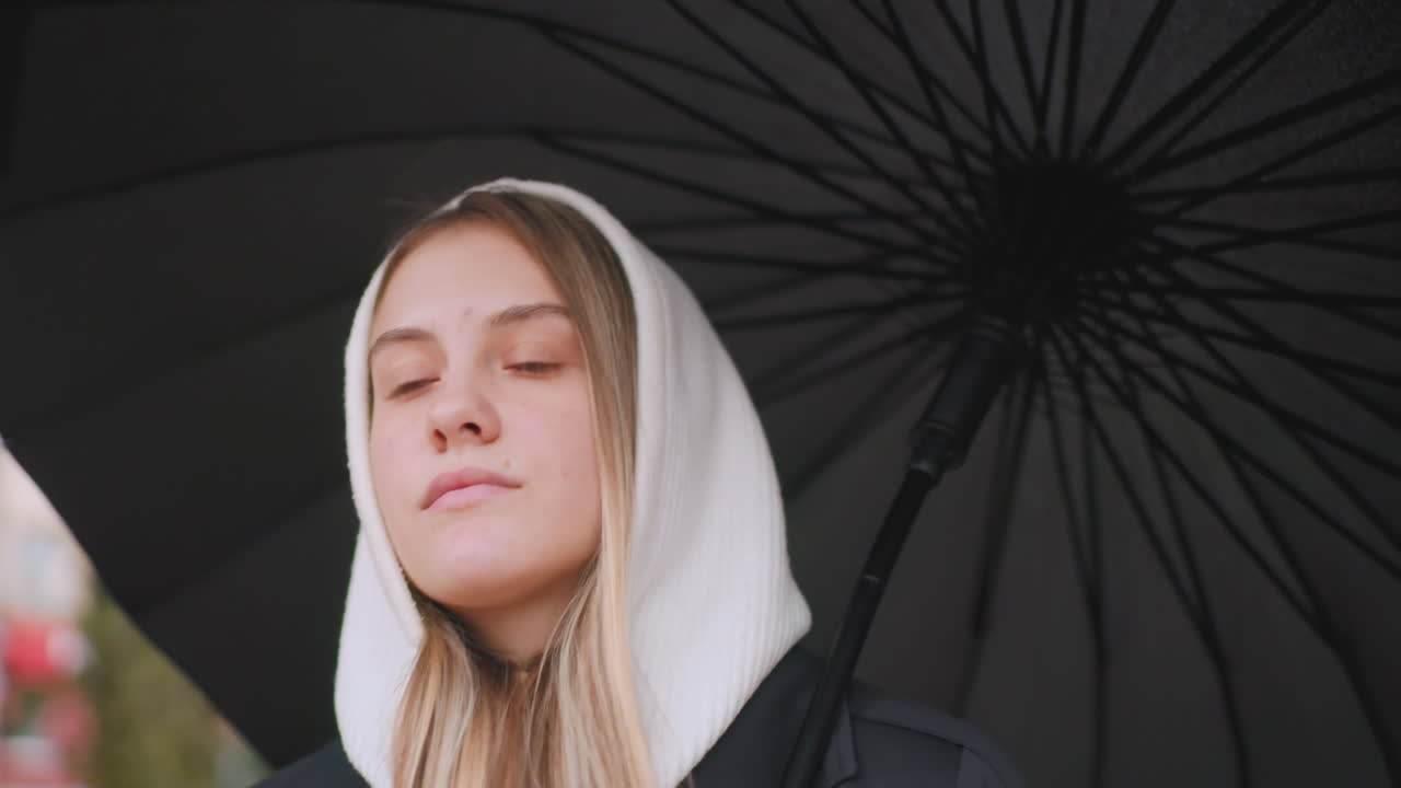 Closeup of young woman in white hood under black umbrella, gazing forward with calm thoughtful expression, natural light highlighting skin and hair, autumn rainy atmosphere, moody